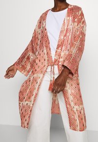 Lätt, mönstrad kimono i nyanser av korall och guld. Har en öppen front, breda ärmar och fransdetaljer.