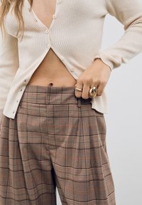 Cardigan crème côtelé avec boutons sur un pantalon à carreaux marron présentant des accents orange. Le modèle montre des bagues superposées sur une main.