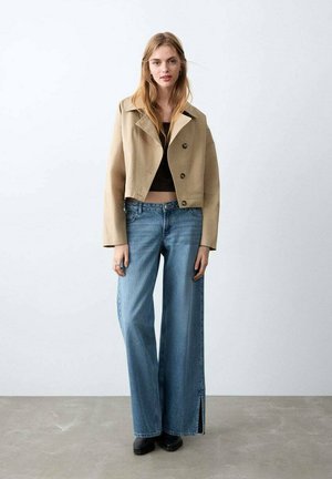 Chaqueta beige recortada con grandes botones, combinada con vaqueros de pierna ancha en azul claro con aberturas en los laterales, llevada con botas negras.