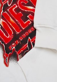 Manica di una felpa bianca che copre parzialmente un testo grafico rosso e nero che recita "DIES" e "FOR SUCCESS" su un fondo bianco.