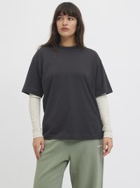 Schwarzes kurzärmliges T-Shirt aus Baumwolle mit lockerer Passform, über einem hellgrauen langärmeligen Oberteil getragen, kombiniert mit grünen Hosen.