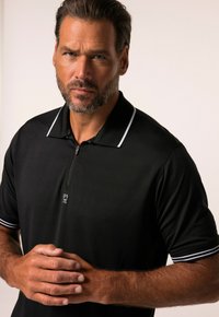 Polo shirt noir à manches courtes avec col zippé, accents blancs et détail de logo. Tissu lisse avec une coupe légèrement ajustée.