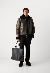 Giacca di pelle marrone con colletto in pelliccia nera, zip frontale e tasche laterali, abbinata a una borsa tote nera in pelle e pantaloni neri.