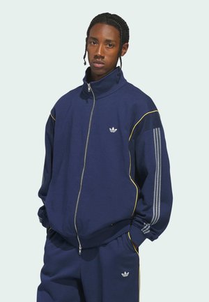 Marineblauw trainingspak gemaakt van zachte stof. Heeft een hoge hals, gele accenten en drie witte strepen op de mouwen en broek. Logo op de borst.