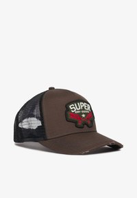 Gorra de tela marrón con lados de malla, visera curvada y un parche bordado de "SUPER DRY GOODS" en verde y rojo. Superficie texturizada con acentos cosidos.