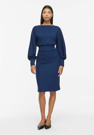 Un mannequin se tient devant un fond blanc uni, portant une robe bleu marine avec une jupe ajustée et de longues manches bouffantes, assortie à des chaussures à talons noires.