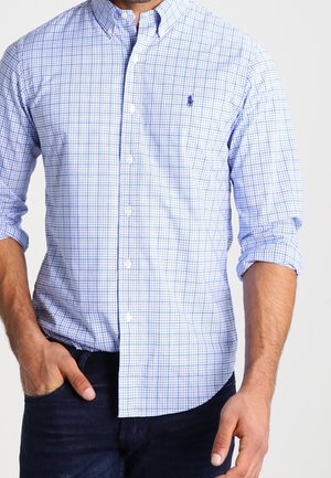 Homme portant une chemise à manches longues bleue et blanche à carreaux avec les poignets retroussés et un jean bleu foncé, main dans la poche.