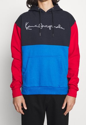 Osoba nosící mikinu s barevnými bloky, černým vrškem, červenými rukávy, modrou střední částí a bílým kurzívním nápisem přes hrudník.