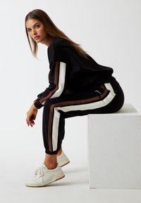 Tuta nera con strisce verticali bianche e marroni, caratterizzata da una vestibilità comoda. Indossata con sneakers bianche, esposta su uno sfondo semplice.