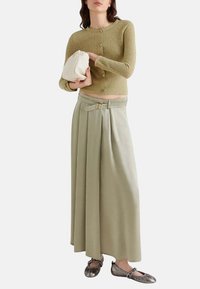 Cardigan verde chiaro a trama con chiusura a bottoni, abbinato a una gonna fluida a vita alta verde chiaro con cintura e pieghe. Décolleté metallici.
