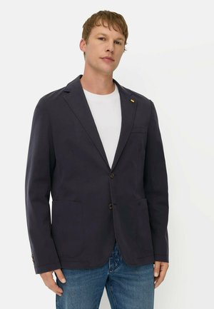 Mand iført en mørk blazer over en hvid skjorte og blå jeans, stående mod en ensartet lys baggrund.