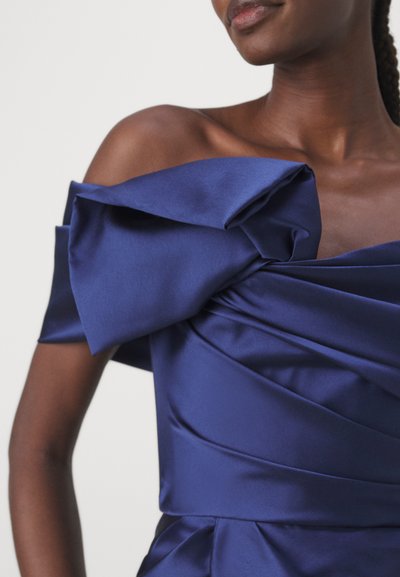 Navy satin dress with off-the-shoulder design, featuring large, draped bows and textured pleats on the bodice. Smooth, glossy finish.