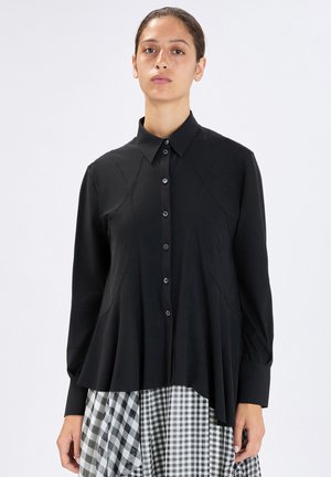 Donna con capelli castani raccolti indossa una camicetta nera con bottoni e motivo ricamato, e una gonna a quadri bianca e nera, sfondo neutro.