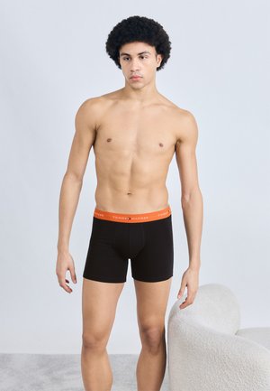 Czarne bokserki z pomarańczowym pasem z napisem "TOMMY HILFIGER" w białym kolorze. Na neutralnym tle.