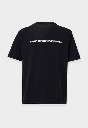Νέος άνδρας στέκεται φορώντας μαύρο T-shirt Emporio Armani, φαρδιά μπλε τζιν και μαύρα αθλητικά παπούτσια σε απλό ανοιχτό φόντο.
