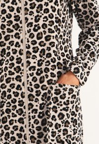 Leopardmönstrad zip-up jacka i mjukt material, med långa ärmar, en framficka och kontrasterande svarta och tan fläckar på en krämfärgad bakgrund.