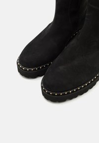 Μαύρες σουέτ μποτες μέχρι τον αστράγαλο με λεία υφή, στρογγυλή μύτη και χρυσές μεταλλικές ακτίνες κατά μήκος της σόλας. Χοντρή, υφασμένη, μαύρη καουτσούκ εξωτερική σόλα.