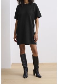 Femme portant une robe courte noire à manches courtes et des bottes en cuir noir montant jusqu'aux genoux, debout sur un sol en bois clair contre un mur blanc uni.