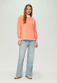 Perzik sweatshirt met gegraveerde tekst, relaxed fit, lange mouwen. Gecombineerd met lichtblauwe wijde jeans en crèmekleurige schoenen.