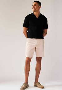 Polo negro de manga corta con cuello en V, combinado con un short de color rosa claro. Lleva zapatos de cordones color tostado, mostrando un atuendo casual de verano.
