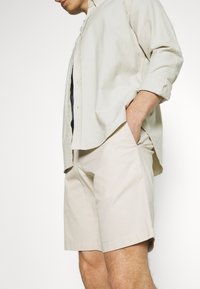 Vaalea beige nappi-paitapusero yhdistettynä matching vaaleanbeigeihin sortseihin, esittäen rentoa istuvuutta ja sileää puuvillakudosta. Ei näkyviä kuvioita tai yksityiskohtia.