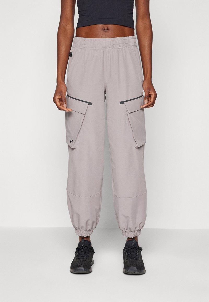 Femme portant un pantalon cargo gris clair avec de grandes poches et des poignets élastiques, un top court noir et des baskets noires, debout sur un fond blanc.
