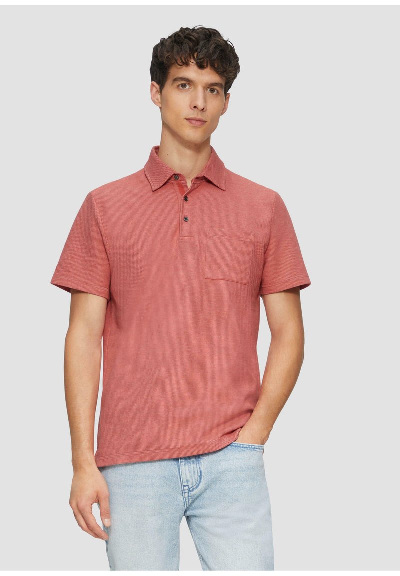 Kurzärmliges Polohemd in einem strukturierten rötlichen Farbton, mit einem Kragen, einer drei-Knopf-Leiste und einer Brusttasche. Kombiniert mit hellblauen Jeans.