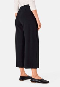 Zwarte brede broek met een gladde textuur en geplooid ontwerp, gecombineerd met zwarte platte schoenen met een lage hak.