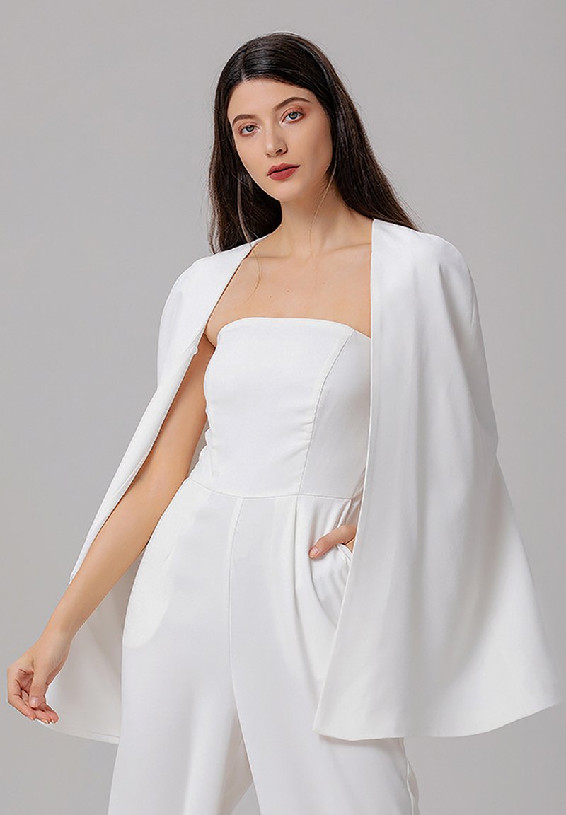 Combinaison en satin blanc avec un design sans bretelles, dotée d'une superposition de cape fluide, de poches latérales et d'une texture lisse et brillante.