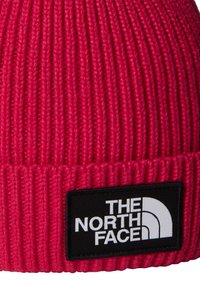 Punainen neulottu pipo, jossa on resorimainen rakenne ja musta suorakulmainen merkki, jossa lukee valkoisella "THE NORTH FACE".