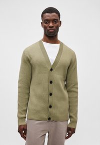 Olivovo zelený rebrovaný kardigán s hlbokým V-výstrihom, piatimi čiernymi gombíkmi a dlhými rukávmi, preložený cez bielu košeľu.