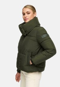 Woman wearing a dark green puffer jacket with a high collar and side pockets, paired with light-colored pants, standing against a plain background.
