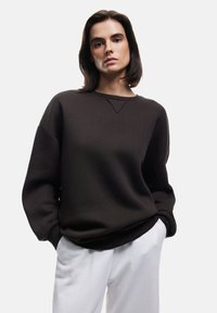 Schwarzes Sweatshirt mit rundem Hals und überschnittenen Schultern, ausgestattet mit gerippten Bündchen und Saum, getragen mit weißen Hosen. Glatte Stofftextur.