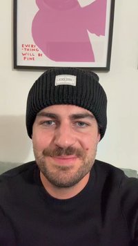 Căciulă tricotată neagră cu o etichetă, purtată de o persoană cu un ten deschis și păr scurt, îmbrăcată într-un pulover închis, pe un fundal neutru.