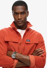 Oranje corduroy overhemd met knopen, twee voorkruiszakken en een label. Witte t-shirt eronder. Zwarte leren horloge om de pols.