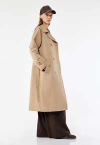 Trench beige con cierre de doble botón, amplios solapas, bolsillos laterales y cinturón de lazo, combinado con pantalones a rayas marrones y una gorra oscura.