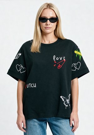 Femme blonde portant des lunettes de soleil noires et un t-shirt noir oversize avec des graphiques colorés et du texte, associé à un jean bleu.
