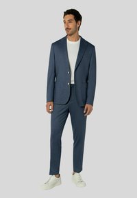 Marineblauer, maßgeschneiderter Anzug mit strukturierter Oberfläche, ausgestattet mit einem Reverskragen, zwei Vorderentaschen und kombiniert mit einem weißen T-Shirt und weißen Sneakern.