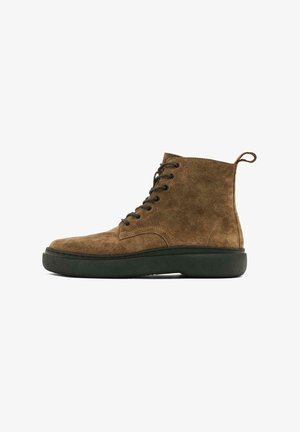 Braune Wildleder-Ankle Boots mit schwarzen Ösen und Schnürsenkeln, runder Zehenpartie und einer grünen Gummisohle. Eine Lasche zum einfachen Anziehen ist vorhanden.
