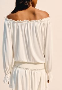 Bílá halenka bez ramínek s volánkovým výstřihem, dlouhými rukávy a staženými manžetami. Obsahuje přiléhající pás s dekorativními akcenty v pase.