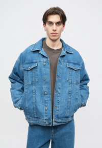 Jaqueta de denim azul com um ajuste descontraído, dois bolsos no peito, botões prateados e um colarinho clássico. Usada sobre uma camisa escura.