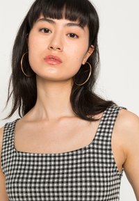 Top com padrão de gingham em preto e branco, com decote quadrado, alças largas e textura suave. Modelo usa grandes brincos de argola dourada.
