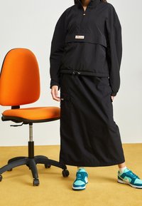 Czarna bluza z pół zamkiem i logo; dopasowana długa spódnica. Modelka nosi turkusowo-białe sneakersy. Pomarańczowe krzesło w tle.