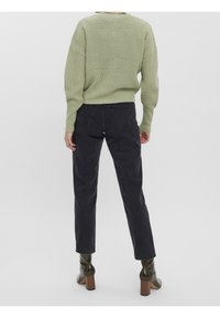 Osoba w jasnzie zielonym swetrze z dzianiny, czarnych dżinsach i ciemnozielonych botkach na obcasie stoi tyłem na jednolitym tle.