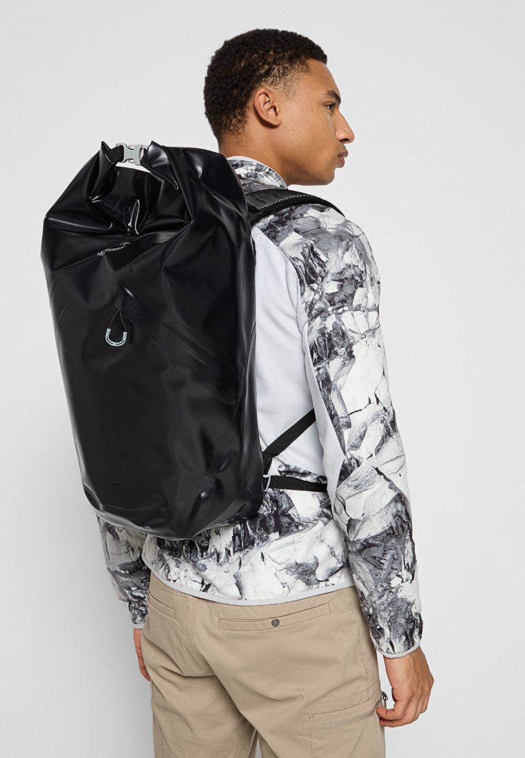 Sac à dos noir étanche avec une fermeture enroulable, des bretelles ajustables et une surface lisse et brillante. Présente un détail de logo de la marque subtil.