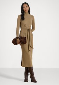 Woman stands wearing a camel turtleneck midi dress with side slit and tie detail, holding a large brown clutch and wearing dark brown boots.