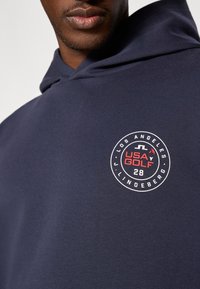 Marineblå hettegenser med et rundt trykt logo som har "USA GOLF," "LOS ANGELES," og "J. LINDEBERG" plassert på brystet. Myk stofftekstur.