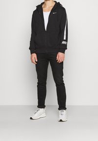 Svart zip-up hoodie med fodrad huva, Nike-logotyp på bröstet och vita detaljer på ärmarna, parat med svarta jeans och vita sneakers.