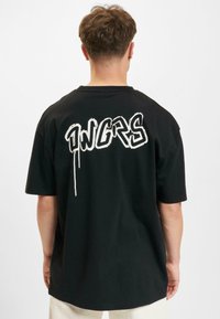 Juodas medvilninis marškinėliai trumpomis rankovėmis ir laisvu kirpimu. Ant nugaros matosi ryškus baltas graffiti stiliaus tekstas "DNGRS".