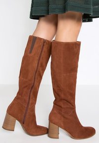 Femme portant des bottes en daim brun à talons blocs et fermeture éclair latérale, assorties à une jupe vert foncé.